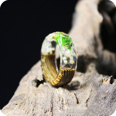 A wood and flower pressed resin cased ring resting upright on a branch. A green and white flower and zebranowood ring