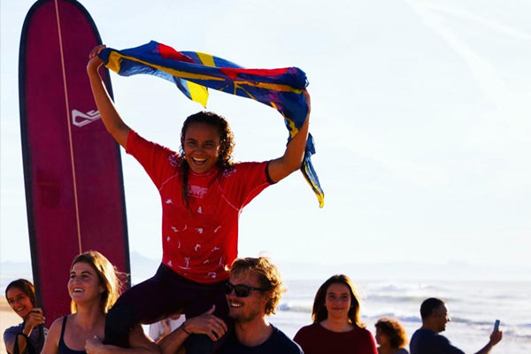 Alice Lemoinge holding country flag whilst being held in the air by friends with surfboard in the background